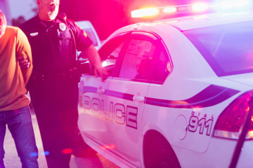 Police officer making an arrest, escorting a young man in handcuffs toward the back seat of his police car, at night.